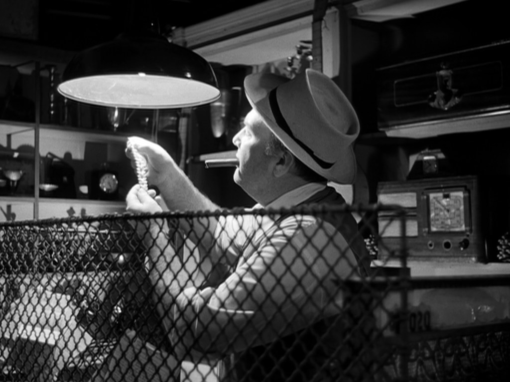A man wearing a fedora inspects an item under a hanging light while surrounded by various objects in a dimly lit room.