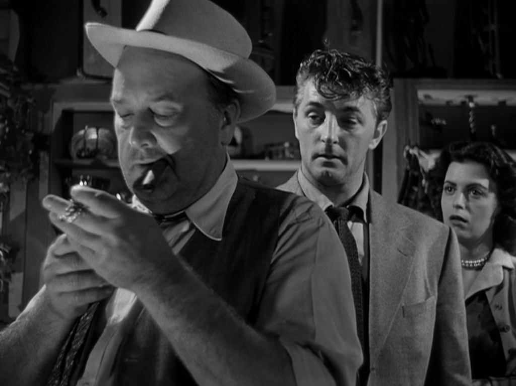 A man in a hat lighting a cigar looks intently, while another man in a suit observes with intrigue, and a woman stands behind them showing concern, in a dimly lit room filled with various items.