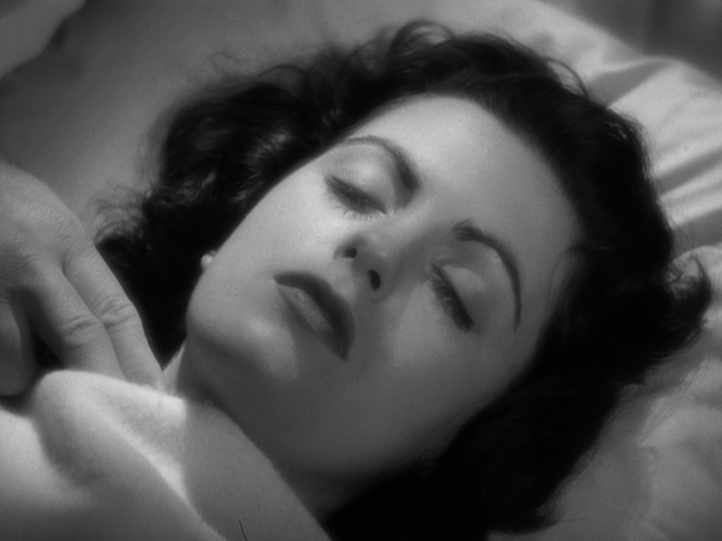 A woman with dark, wavy hair lying peacefully on a bed, eyes closed, suggesting a serene or sleeping state.