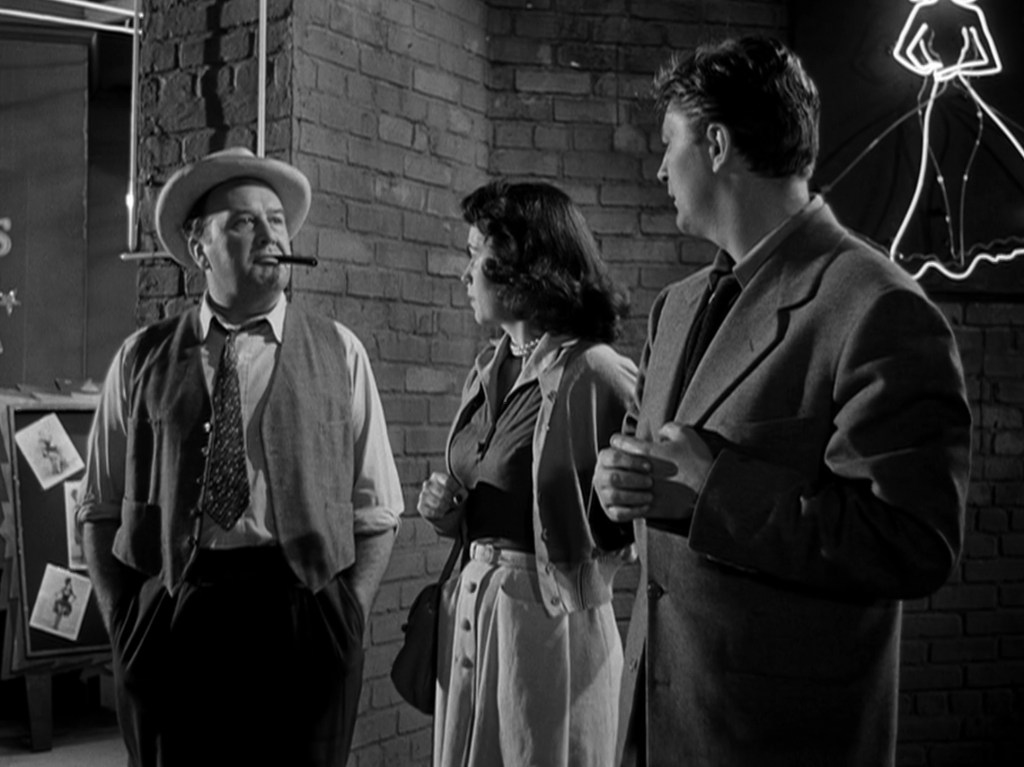 A scene featuring three characters in a black and white setting, with a man wearing a hat and holding a cigar, a woman in a light-colored skirt and dark top, and another man in a suit, engaged in conversation against a brick wall with neon signage.