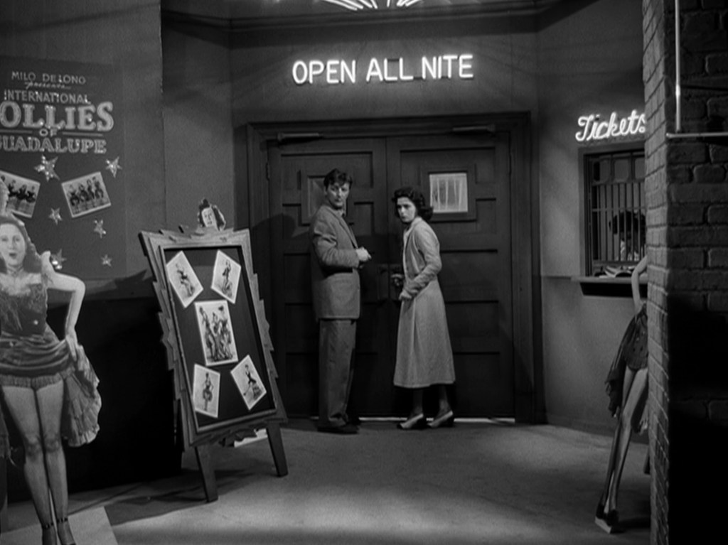 Two people stand in front of a dimly lit entrance with the sign 'OPEN ALL NITE.' Nearby are posters advertising a show and a ticket booth in the background.