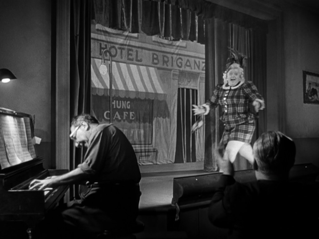 A performer in a plaid costume energetically entertains on stage while a pianist plays, and audience members applaud in the foreground.