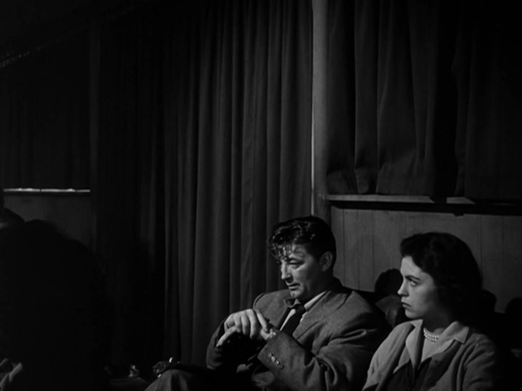 A man and woman sitting side by side in a dimly lit setting, with curtains in the background. The man appears deep in thought, while the woman looks contemplative.