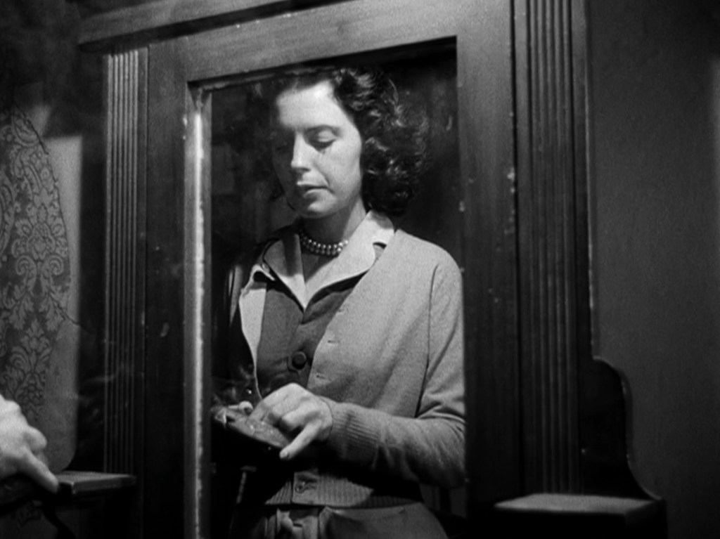 A woman with curly hair stands inside a vintage phone booth, holding a receiver and looking thoughtfully. She is wearing a cardigan and a pearl necklace. The setting appears dark and nostalgic, with intricate wallpaper visible in the background.
