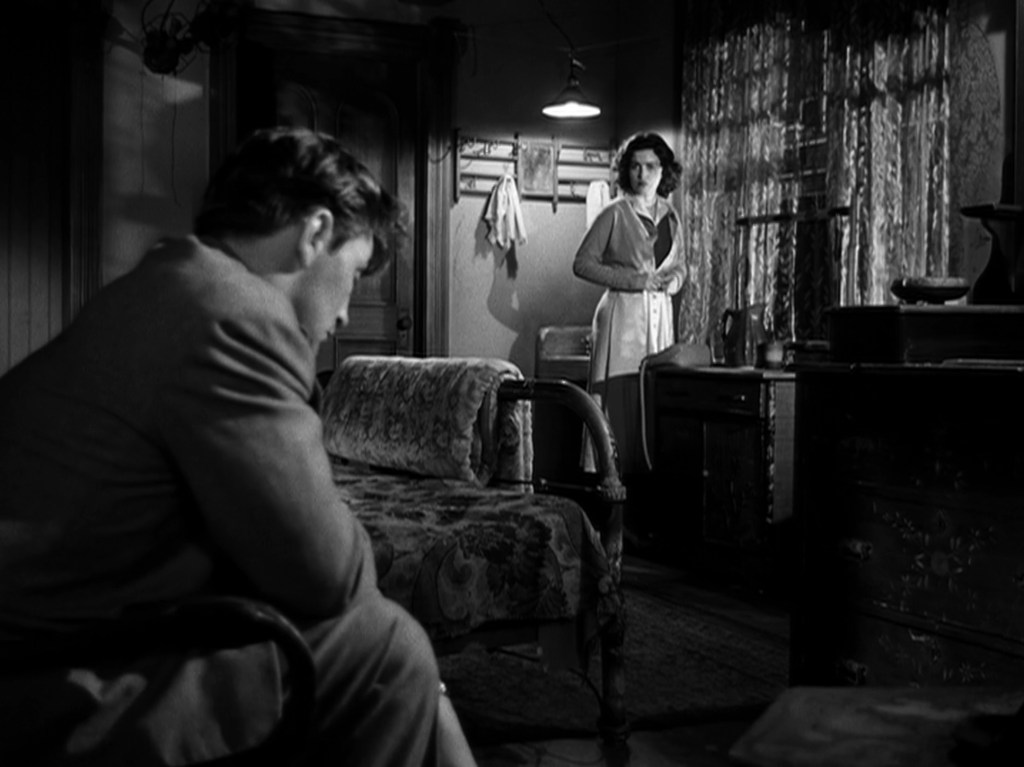 A man seated on a couch, looking down, while a woman stands in a dimly lit room, wearing a light dress and looking at him.