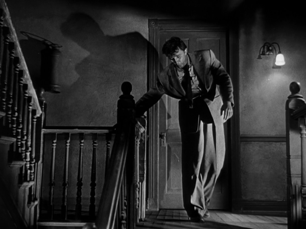 A man in a suit walks down a dimly lit staircase, casting a large shadow on the wall behind him.