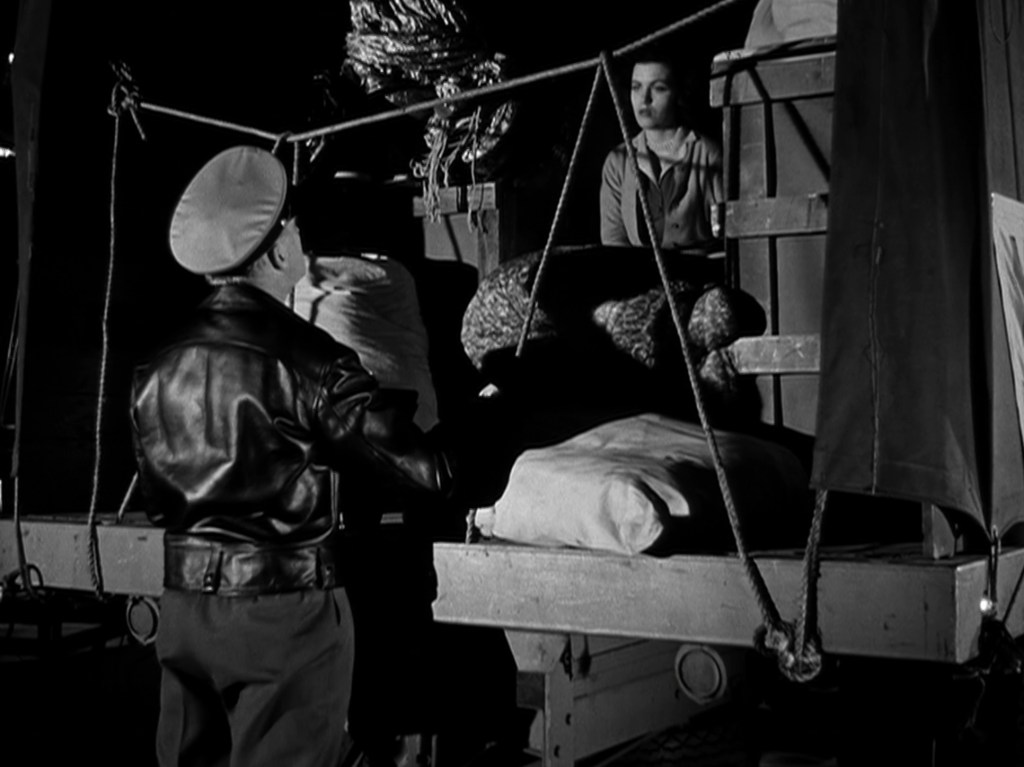 A soldier in a leather jacket and cap interacts with a woman sitting on a stacked cargo in a dimly lit setting.