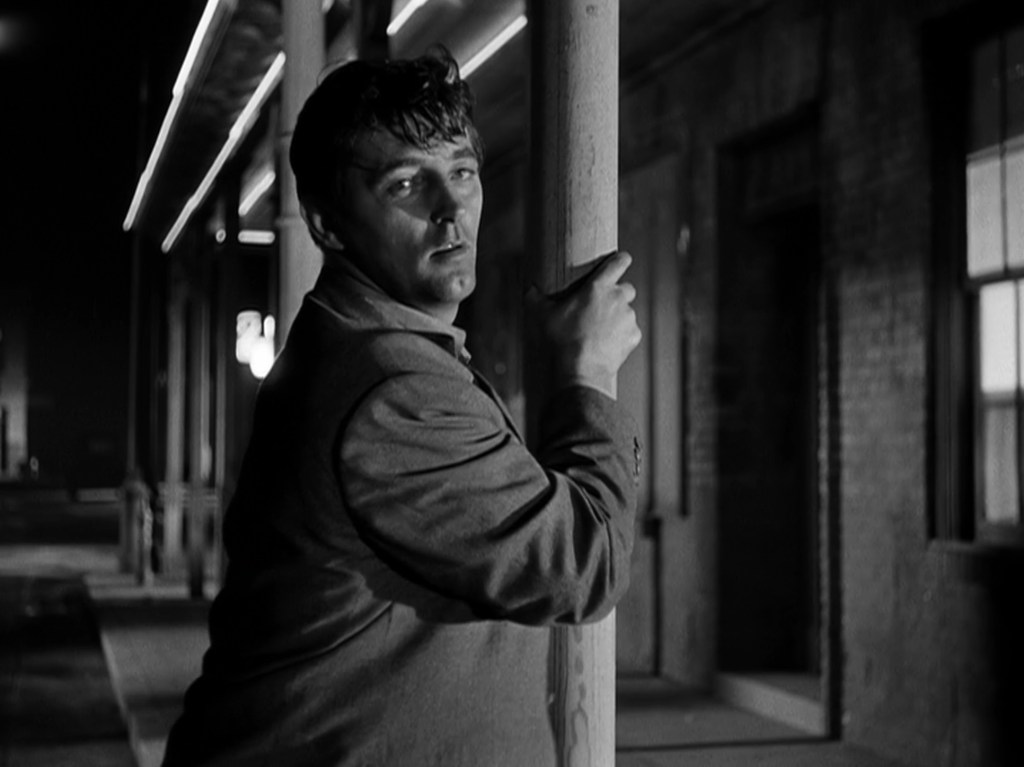 A man in a suit leans against a wooden pole in a dimly lit, urban setting. The scene is in black and white, highlighting his contemplative expression.