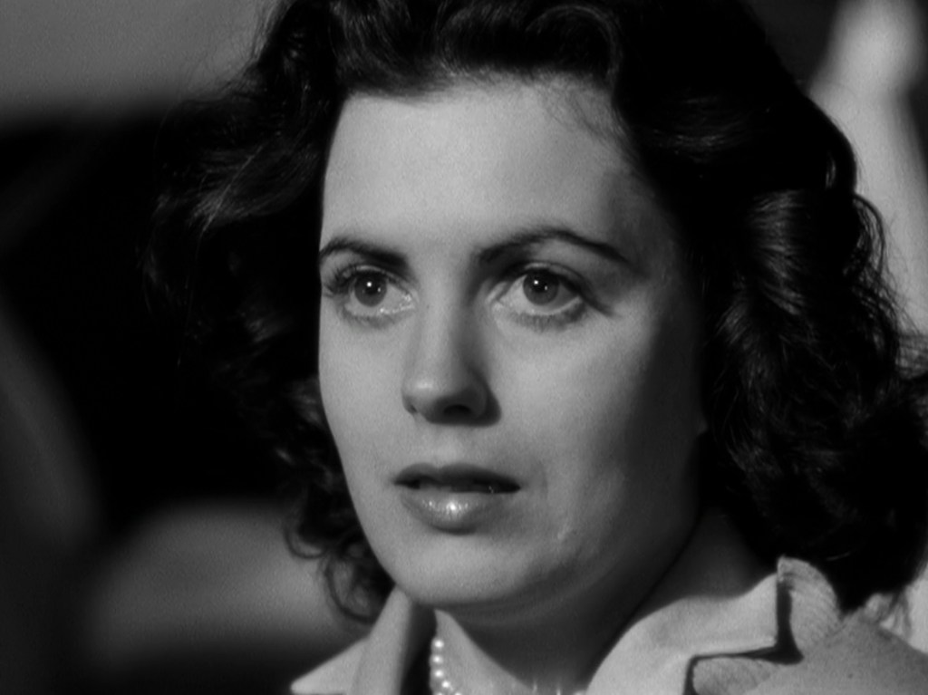 Close-up portrait of a woman with curly hair, wearing a light-colored blouse and a pearl necklace, reflecting a pensive expression.