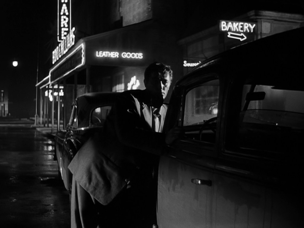 A man in a suit leans against a vintage car at night, with illuminated shop signs in the background, including 'Bakery' and 'Leather Goods.'