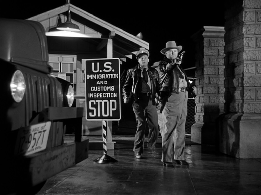 Two officers in uniforms stand next to a 'U.S. Immigration and Customs Inspection STOP' sign, with a parked vehicle in the foreground.