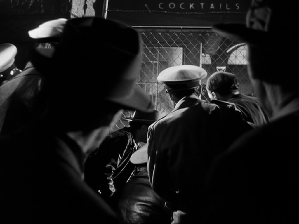 A group of people in hats gathered outside a door labeled 'Cocktails', with a focus on their backs and side profiles.