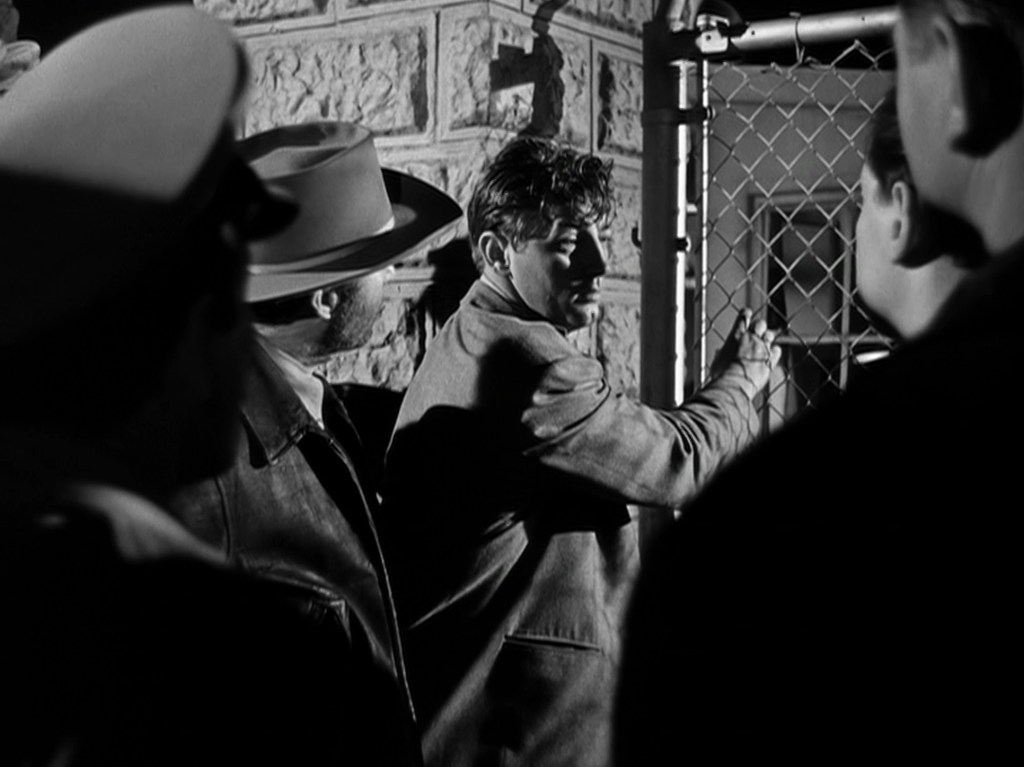 A dramatic black-and-white scene depicting a man reaching for a gate in a tense moment, surrounded by shadowy figures, suggesting a confrontation or secretive exchange.