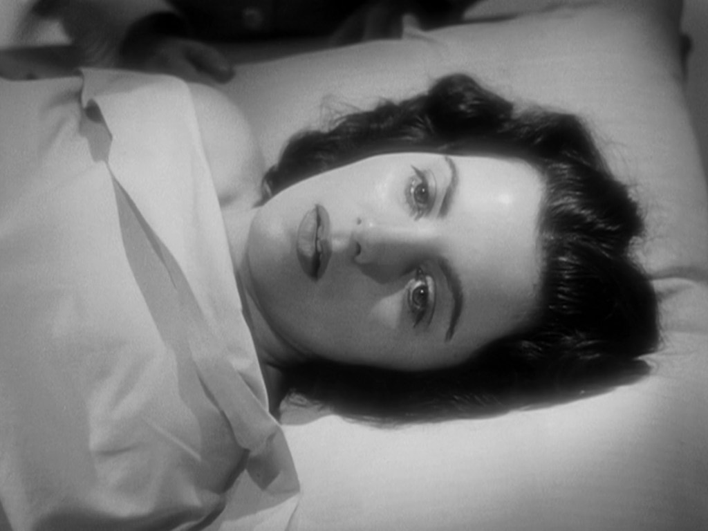 A close-up of a young woman lying on a pillow, looking up with a serene expression, draped in a light-colored sheet.