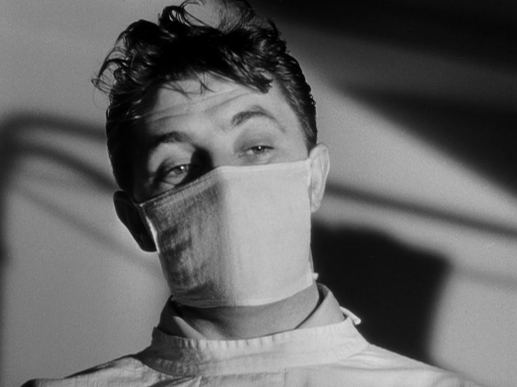 A male figure wearing a surgical mask and a medical gown, gazing intently with a shadowy background.