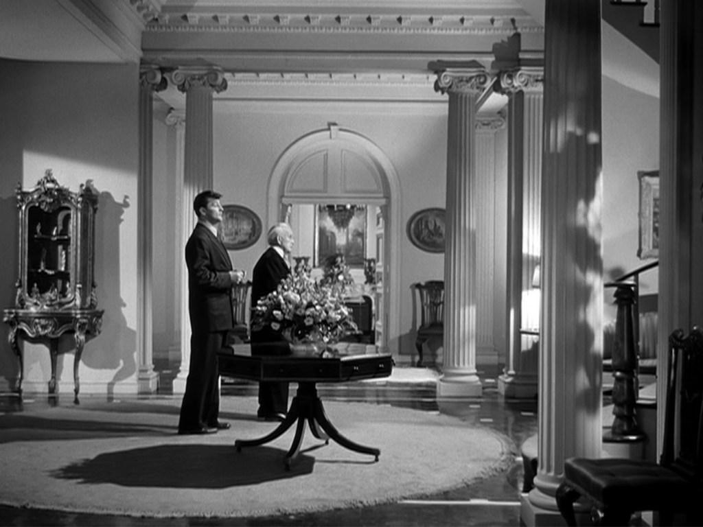 Two men standing in a spacious, elegantly furnished room with tall columns and a large floral arrangement on a table.