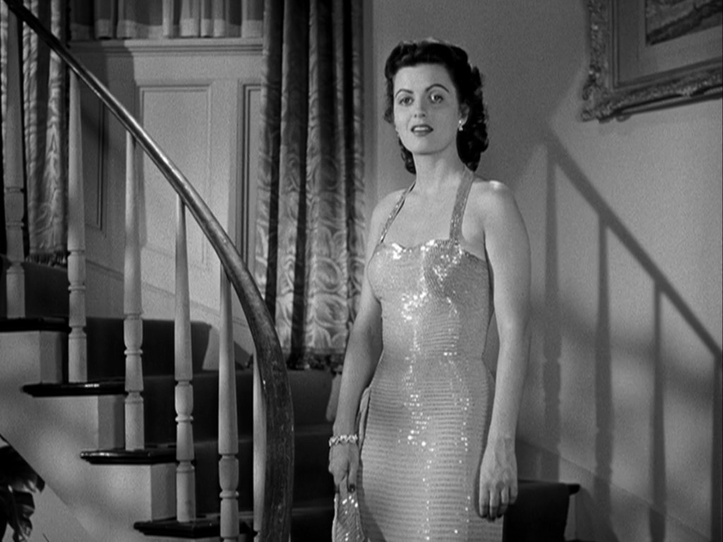 A woman in a sparkly, form-fitting dress stands at the bottom of a staircase, with a poised expression. The background features elegant curtains and decorative wall art.