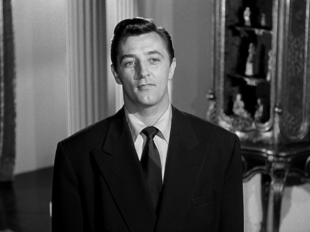 Black and white image of a man in a formal suit, standing in a room with elegant decor, looking slightly to the side.
