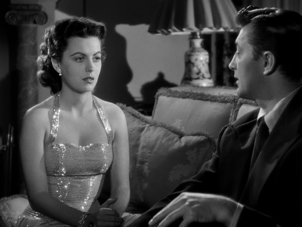 A woman in a sparkling evening gown sits on a couch, looking thoughtfully at a man beside her who is speaking. The setting is dimly lit, with a lamp in the background.