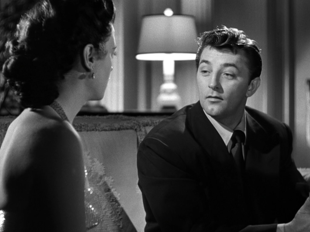 A man in a black suit engages in conversation with a woman, who has her back to the camera. The scene is set in a softly lit room with a lamp in the background.