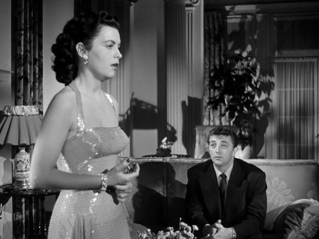 A woman in a sparkling dress stands speaking, while a man in a suit sits nearby, looking contemplative. The scene has a vintage black-and-white aesthetic with elegant furnishings in the background.