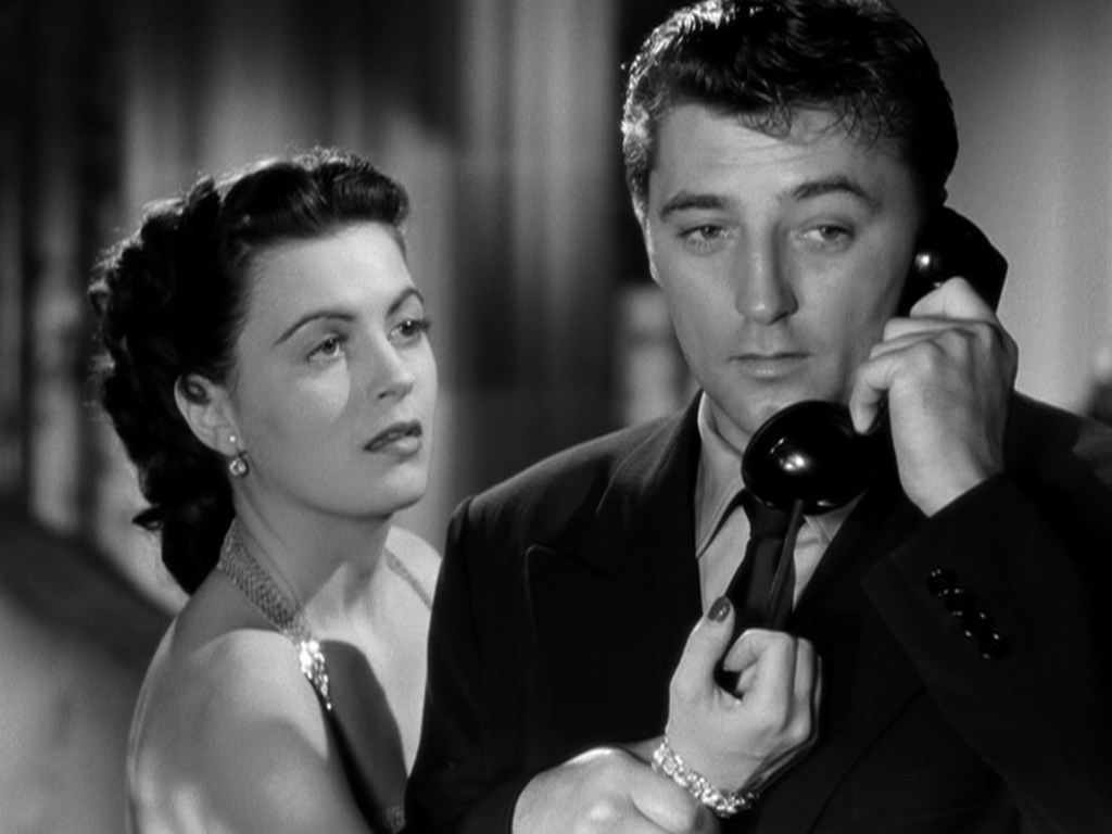 A woman looks intently at a man holding a phone, conveying a sense of tension or concern, in a classic black and white setting.