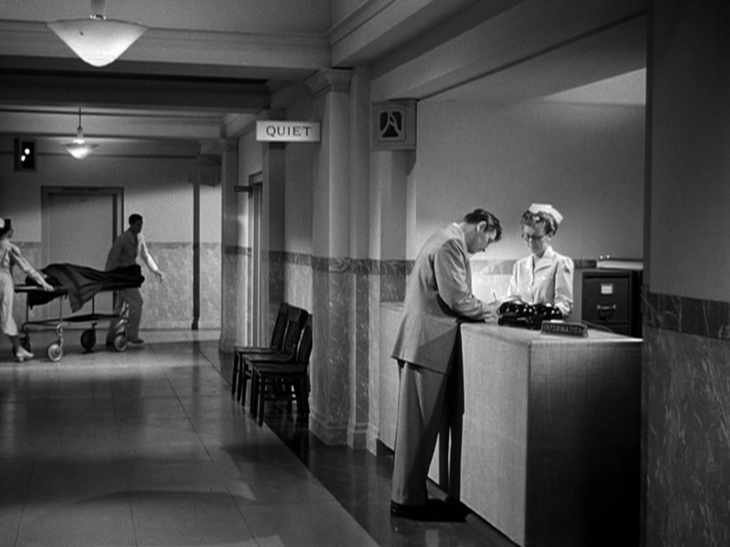 A hospital corridor featuring a nurse at an information desk talking to a man. A second nurse pushes a stretcher in the background. A 'QUIET' sign is visible on the wall.