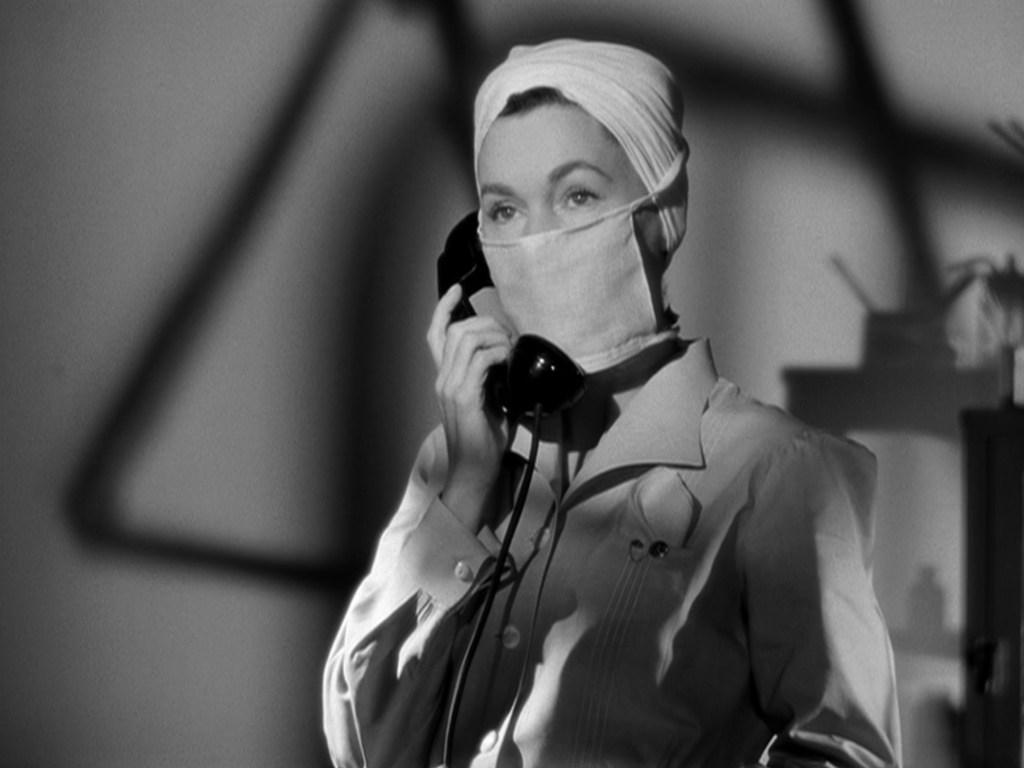 A woman wearing a headscarf and a mask speaks on a vintage telephone in a black and white setting.