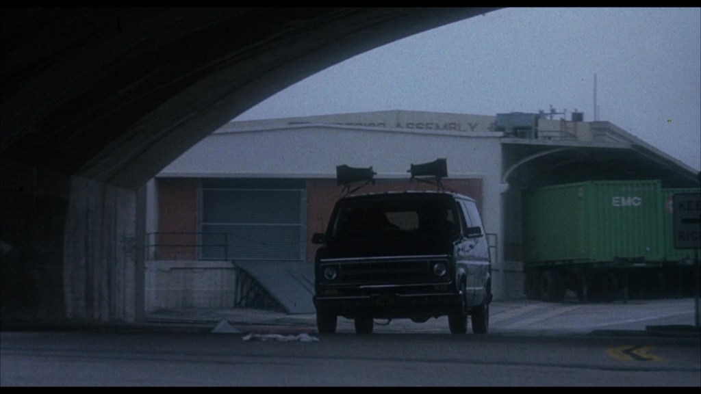 A black van with loudspeakers on top is parked under a bridge, with an industrial building and a green shipping container in the background.