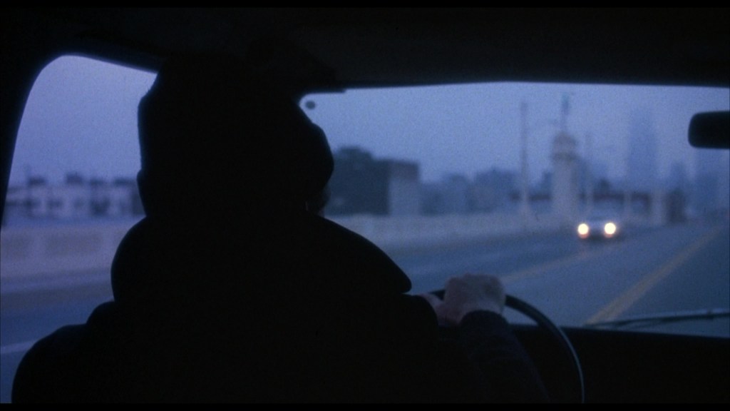 A shadowy figure driving a vehicle at dusk, facing an illuminated street with distant city buildings in the background.