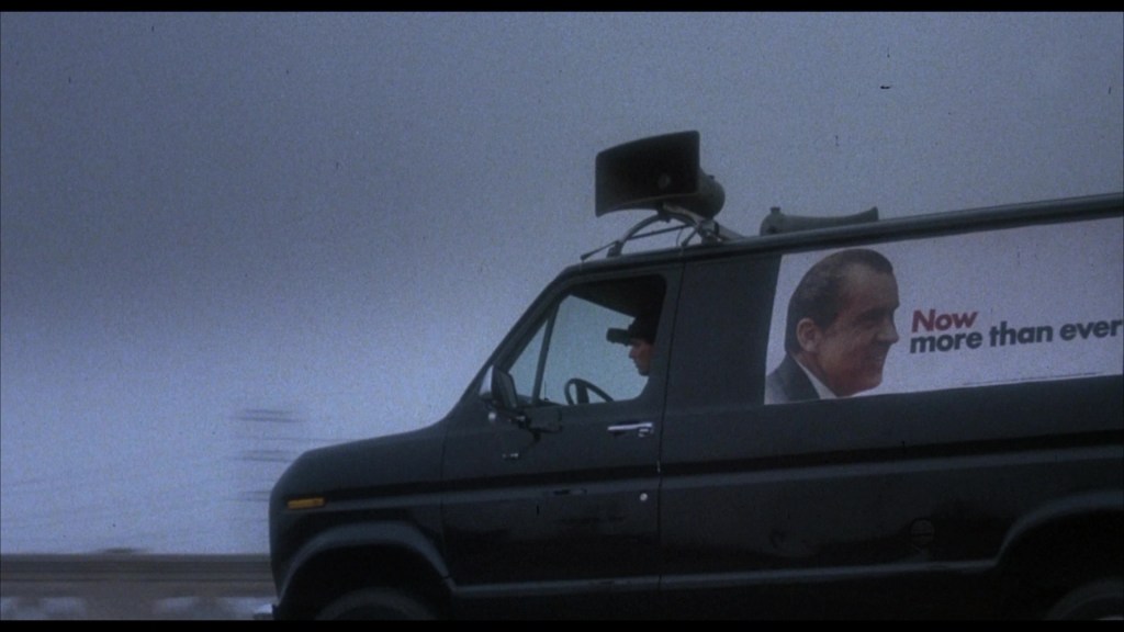 A black van with a large image of a man and the text 'Now more than ever' on the side, driving on a foggy road.
