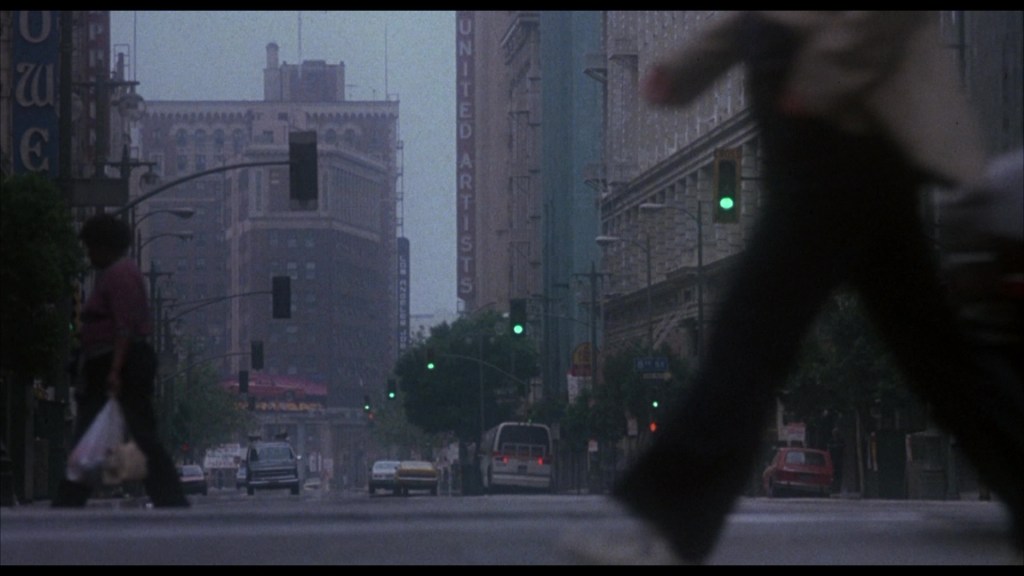 A city street scene with pedestrians walking past traffic signals and buildings, featuring a sign for 'United Artists' in the background on a foggy day.