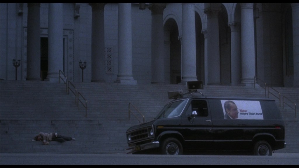 A dark van parked near a set of large stairs outside a building, with one person lying on the ground nearby.