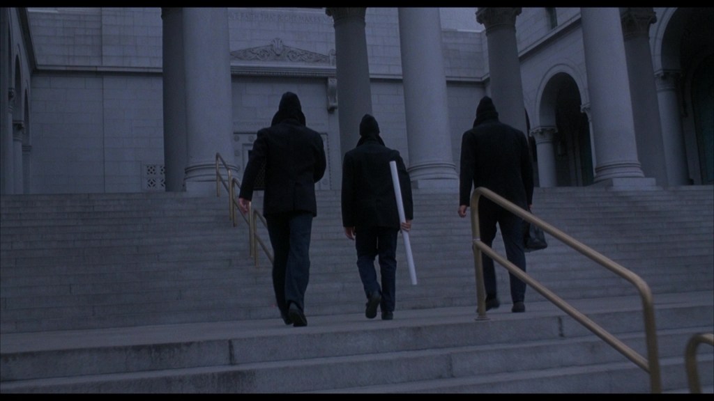 Three figures wearing dark clothing and hooded jackets walk up a set of stone steps beside a railing at a monumental building. One figure carries a large white object.