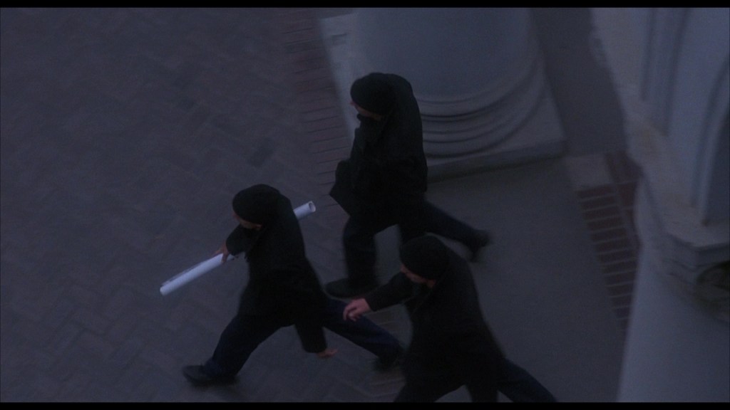 Three individuals dressed in black clothing, walking along a stone pathway, one holding a rolled-up document.