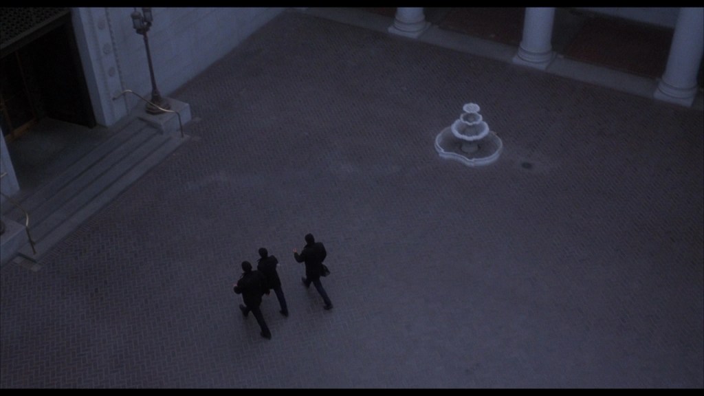 Three figures in black clothing walking towards a fountain in an illuminated outdoor area, with a building entrance visible in the background.