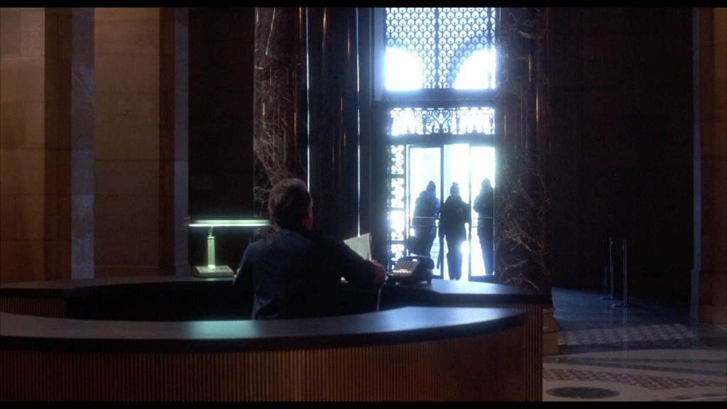 A view of a reception desk with a person sitting on a chair, looking towards a large entrance where silhouettes of three people are entering.