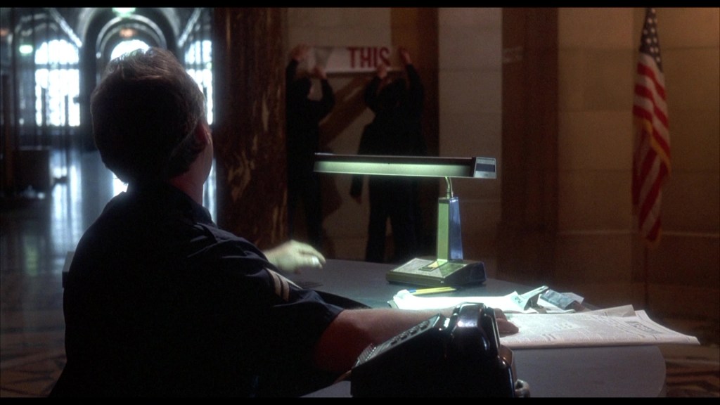 A security guard sits at a desk in a hall, looking towards two individuals in the background who are holding a sign that says 'THIS.' An American flag is visible to the right.