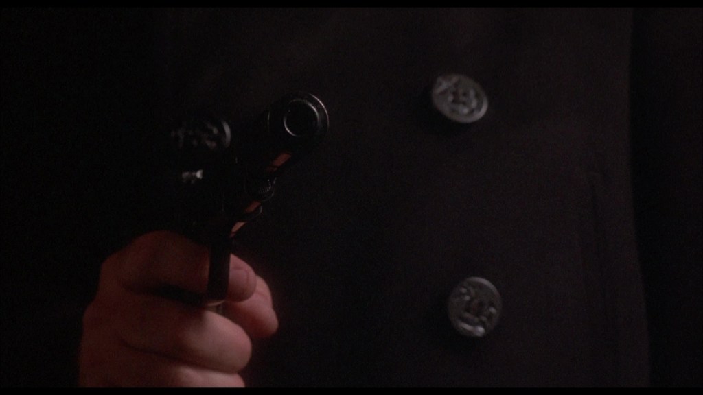 A close-up of a hand holding a pistol, wearing a black coat with visible buttons.