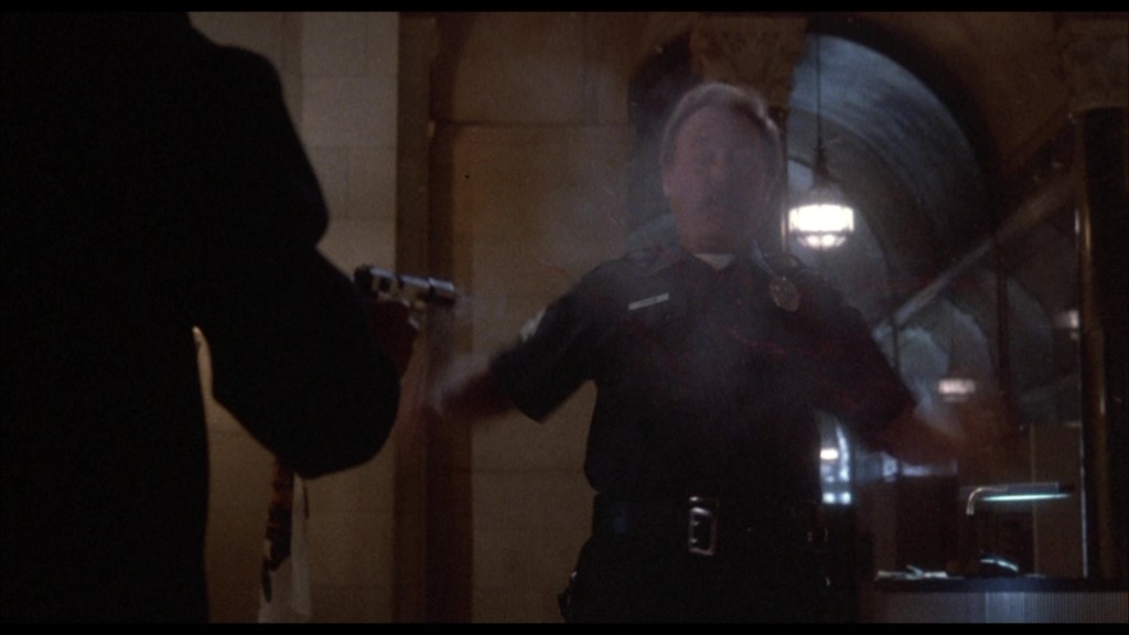 A police officer appears startled as he stands in an indoor setting, with smoke suggesting a recent gunshot. A hand holding a firearm is visible in the foreground.