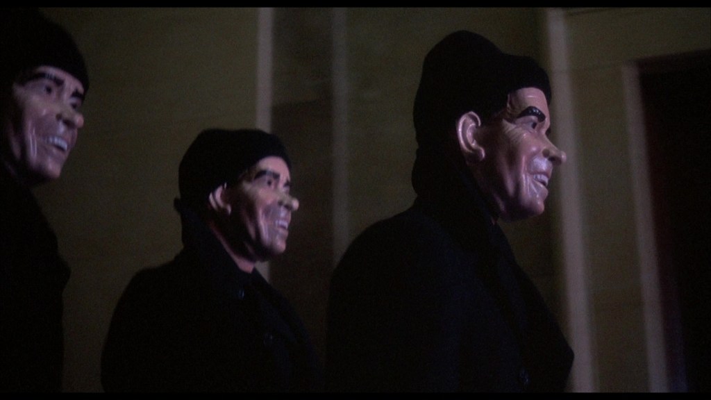 Three figures wearing identical masks and black clothing walking in low light