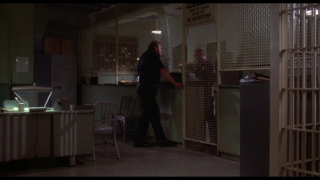 A police officer stands at a gated entrance speaking with another person inside a dimly lit office space, with a desk and chairs visible.