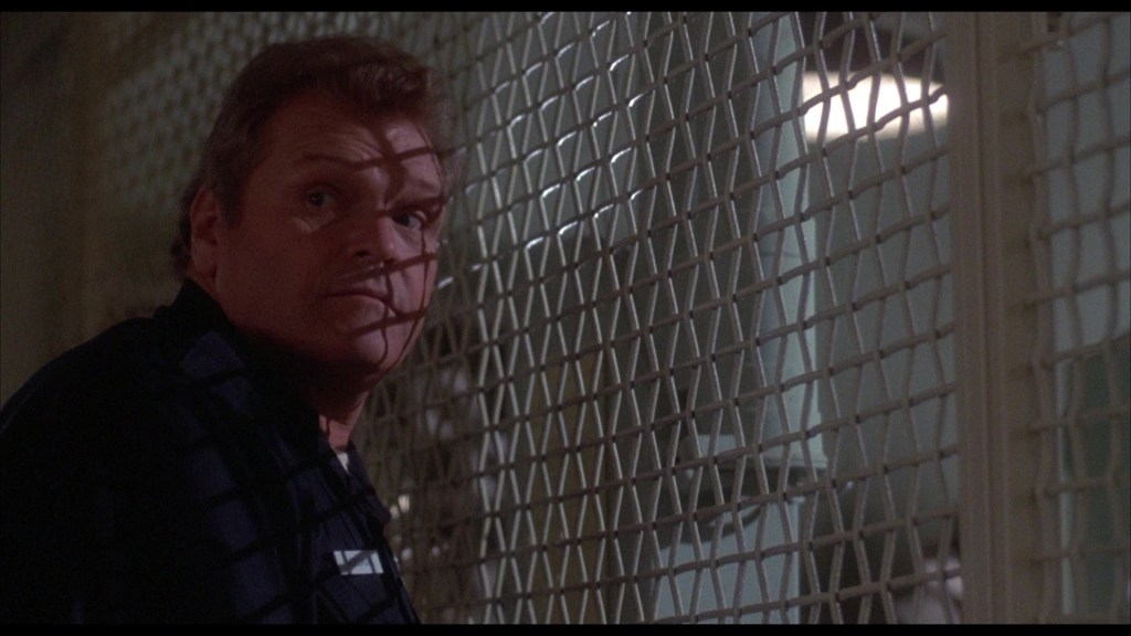 A man in a dark outfit glances over his shoulder, with shadows cast across his face from a nearby grate.