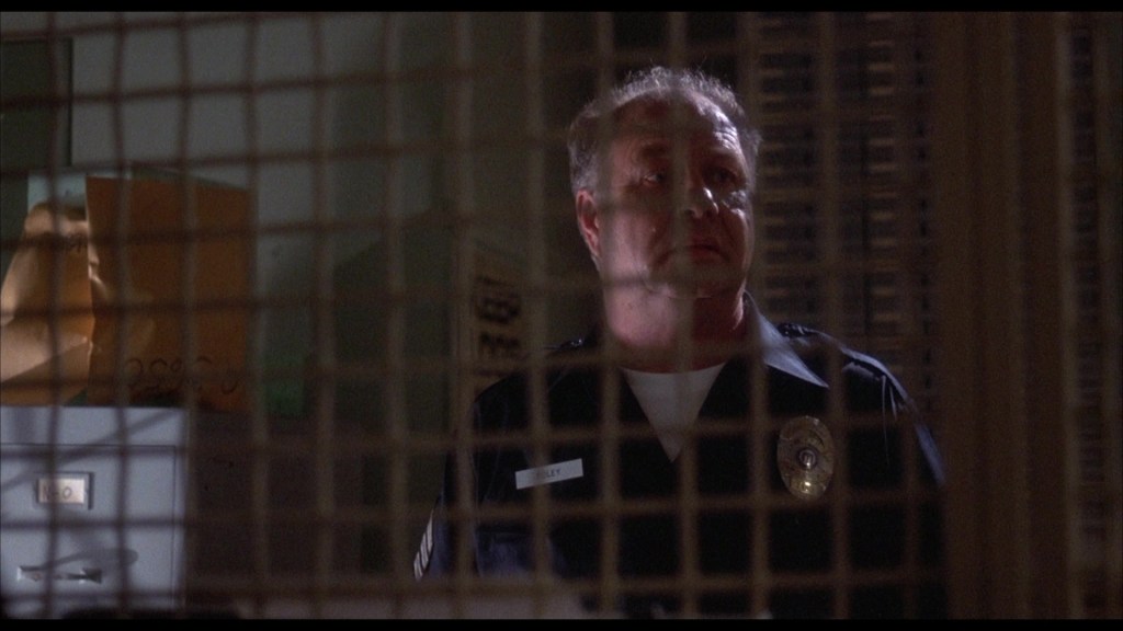 A police officer standing near a file cabinet, visible through a metal grate, with a concerned expression.