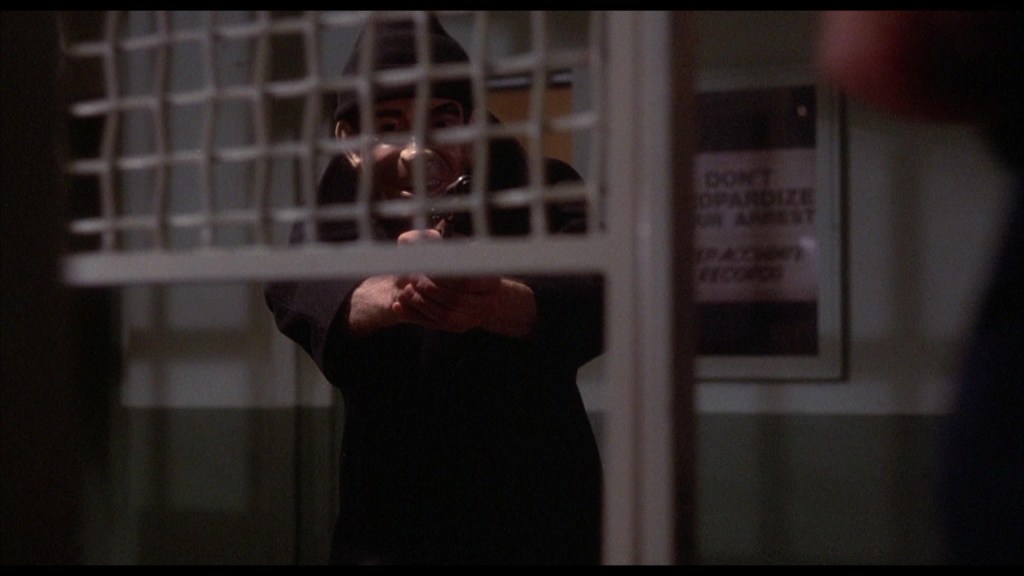 A shadowy figure in a hooded outfit points a handgun through a barred window, creating a tense atmosphere.