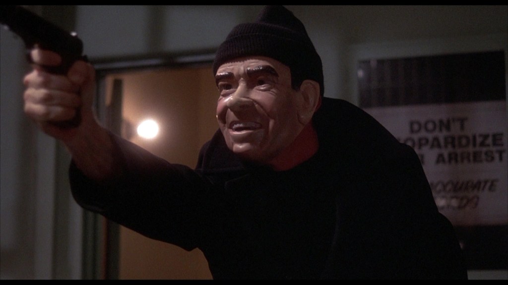 A person wearing a ski mask and a mask resembling a character's face points a gun menacingly in a dimly lit room.