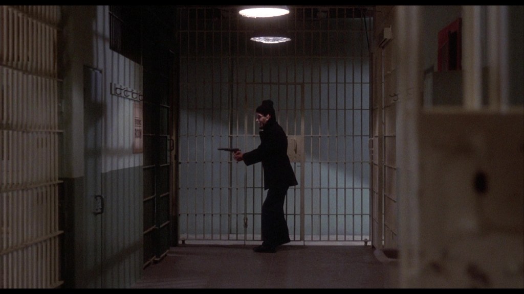 A person wearing a dark outfit and hat stands in a dimly lit prison corridor, holding a gun, with bars visible in the background.