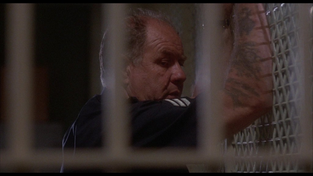 A man with tattoos leans against a barred wall, appearing contemplative in a dimly lit environment.