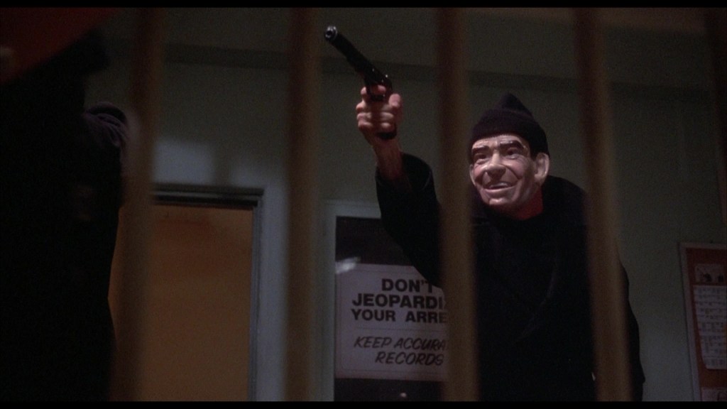 A man wearing a black outfit and a mask holds a gun, aiming it with a serious expression. The background features a sign with text warning not to jeopardize an arrest.