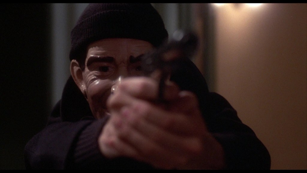A figure wearing a black beanie and dark clothing points a gun directly at the camera, revealing a mask with exaggerated facial features.